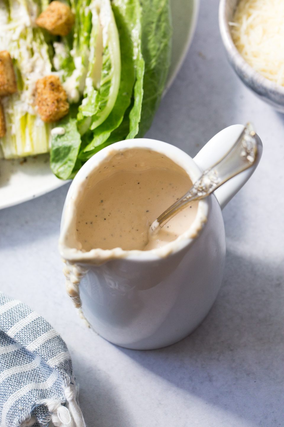 An overhead shot of a white gravy dish with caesar dressing inside. There is a spoon placed inside the container and then caesar salad in the upper left corner. There is a blue striped napkin draped in the lower left corner.