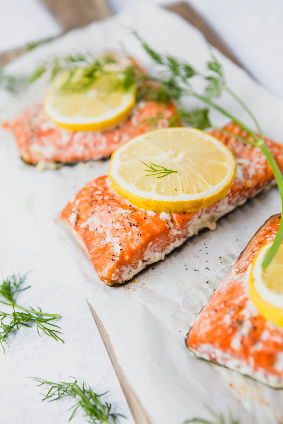 A 45 degree angle shot of three pieces of cooked salmon on parchment paper. Topped with lemon slices and garnished with dill.